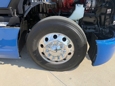 Kenworth T680 front wheel assembly showing polished aluminum hub with multiple lug nuts; blue fender and black tire with tread visible.