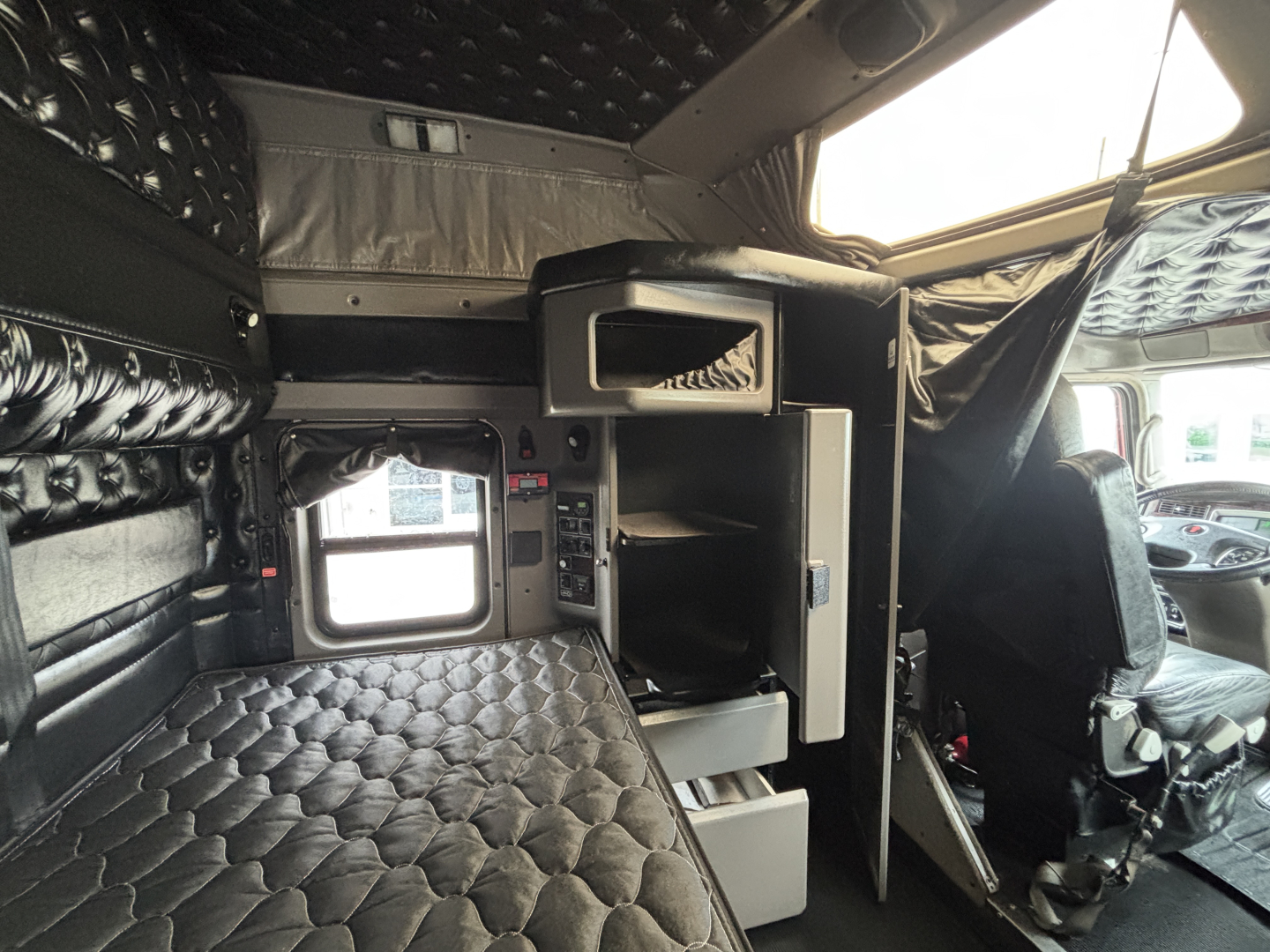Kenworth W900L truck cabin interior with quilted black upholstery and sleeper area, visible storage compartments and driver seat.