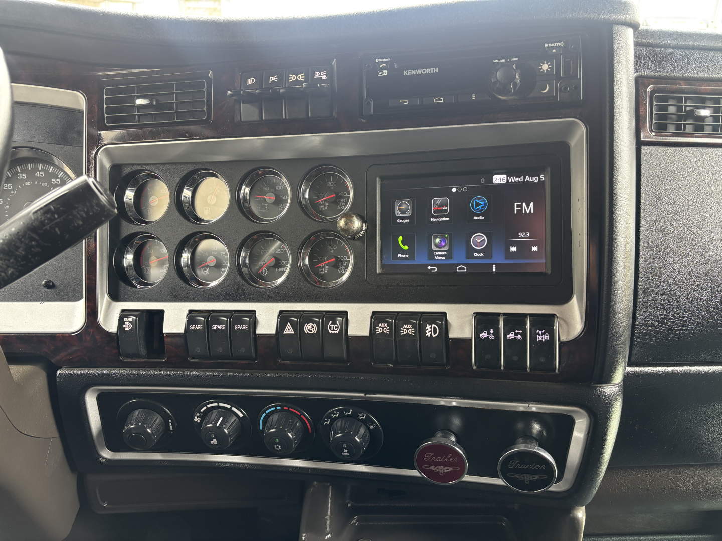Kenworth W900L dash with multiple gauges, central touchscreen display showing FM and apps, row of toggle switches, climate controls, and two round knobs; dashboard chrome trim.