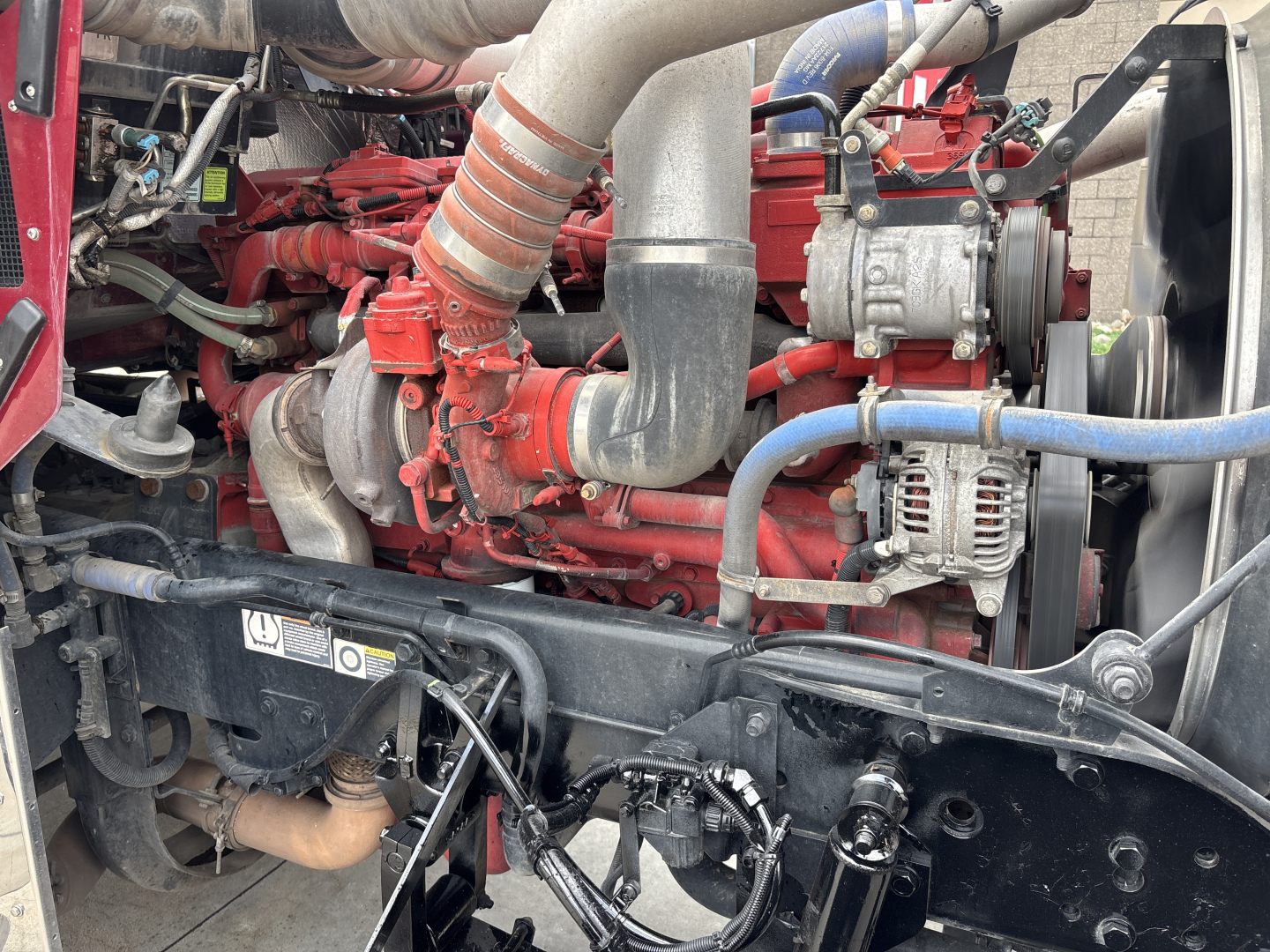 Kenworth W900L engine bay showing red engine components, intake piping, alternator, hoses, and exposed wiring.