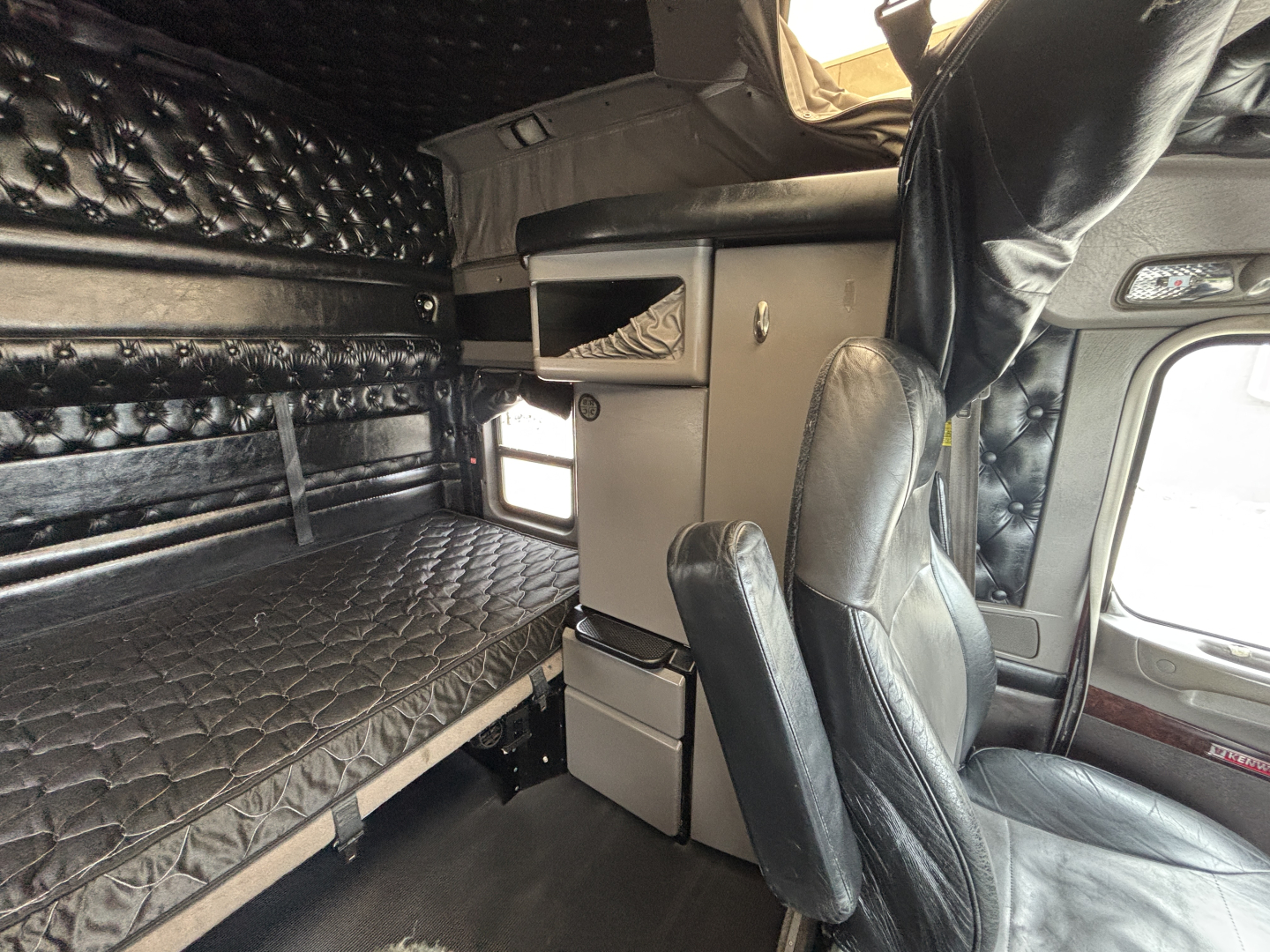 Interior of a Kenworth W900L sleeper cab showing bunk bed with quilted mattress, storage drawers, and driver seat; no legible model text visible.