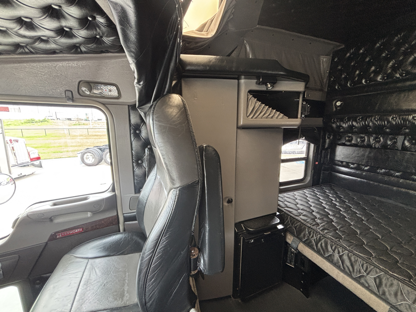 Kenworth W900L truck interior showing sleeper bunk and captain’s chair with black quilted upholstery and storage compartments.