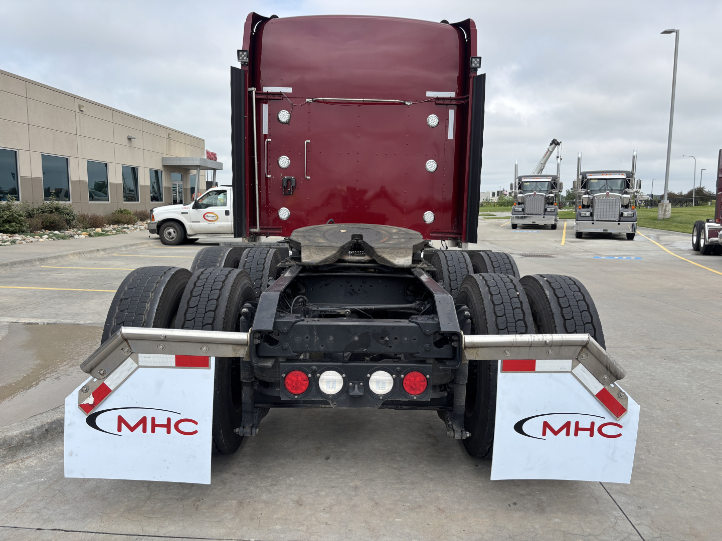 Kenworth W900L rear view showing dual rear axles and mudflaps with MHC logos, red sleeper cab, parking lot.