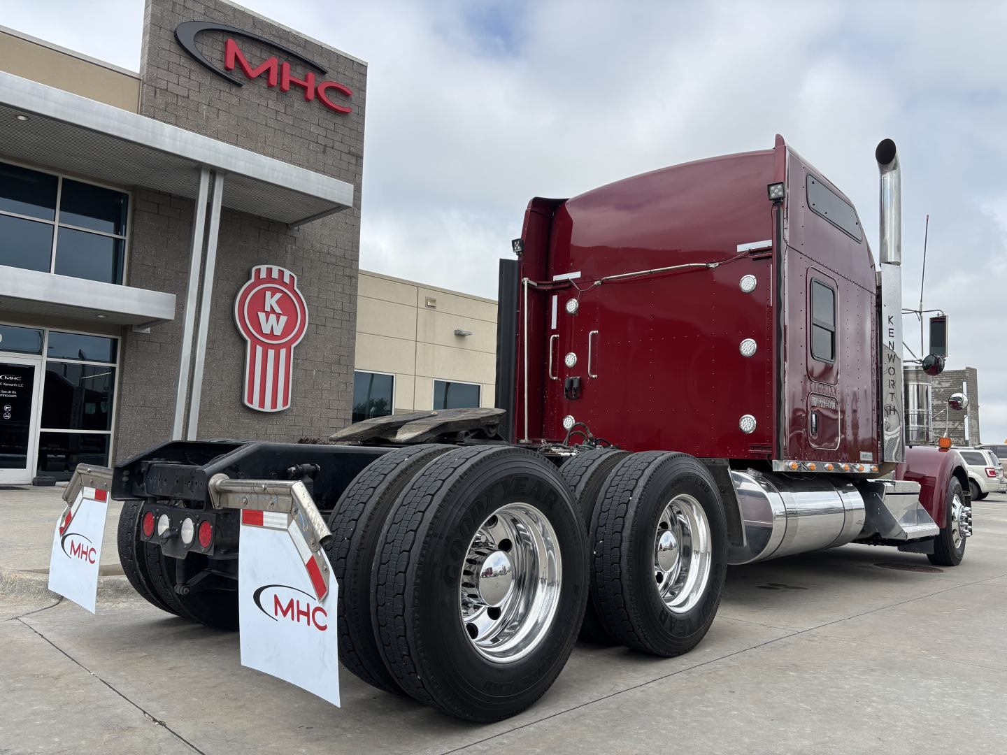 Kenworth W900L semi-truck in red cab, dual rear axles visible, side view showing chrome fuel tanks and exhaust stack, MHC dealership building in background.