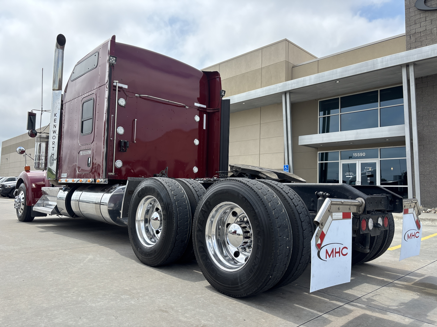 Kenworth W900L tractor-trailer chassis with dual rear axles, maroon cab, chrome fuel tanks, and mudflaps labeled MHC. Visible text includes 'KENWORTH' on the cab and 'MHC' on mudflaps.