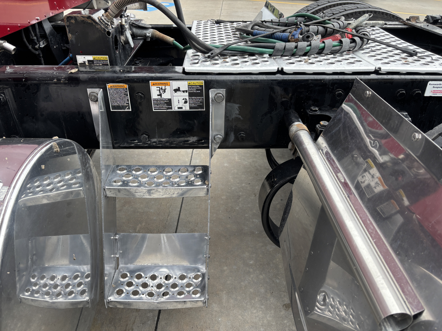 Kenworth W900L undercarriage showing steps, fuel lines, and chassis with access panels and metal tubing.