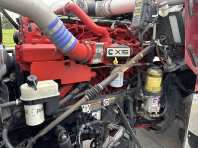 Kenworth W900L engine bay showing red engine block with X15 badge, blue air intake hose, yellow oil filter, and various hydraulic lines.