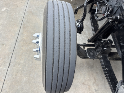 Kenworth W900L front wheel showing dual rear axles and suspension components with a single wide low-profile tire, chrome lug nuts on hub, and chassis frame.