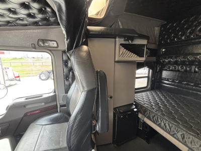 Kenworth W900L truck interior showing sleeper bunk and captain’s chair with black quilted upholstery and storage compartments.