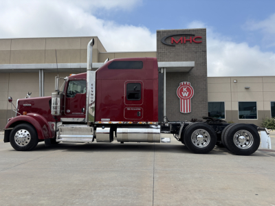 Kenworth W900L truck side view showing tractor cab and dual rear axles at a dealership.