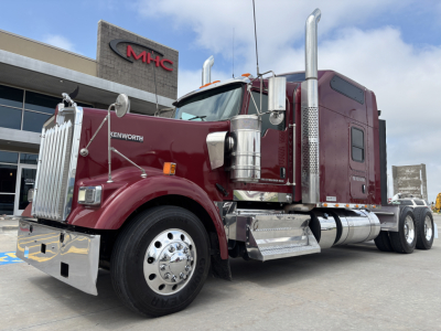 Kenworth W900L truck in maroon color, chrome fuel tanks and exhaust stacks, front grille, headlight on left, large front bumper. Visible text: KENWORTH on the side of the hood. Behind is a dealership building.