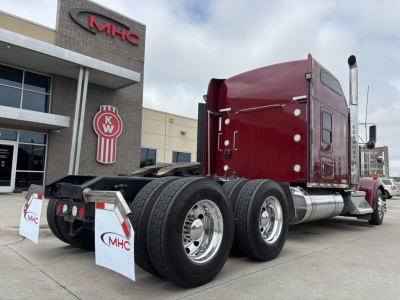 Kenworth W900L semi-truck in red cab, dual rear axles visible, side view showing chrome fuel tanks and exhaust stack, MHC dealership building in background.