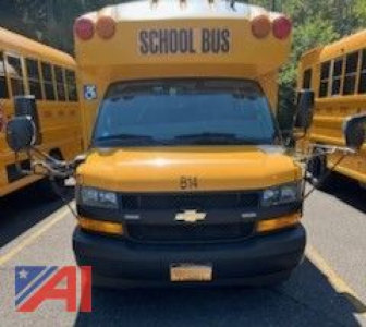 Front view of a 2019 Chevrolet Trans Tech Express mini bus with wheelchair lift, yellow school buses on both sides, Chevrolet emblem on grille.