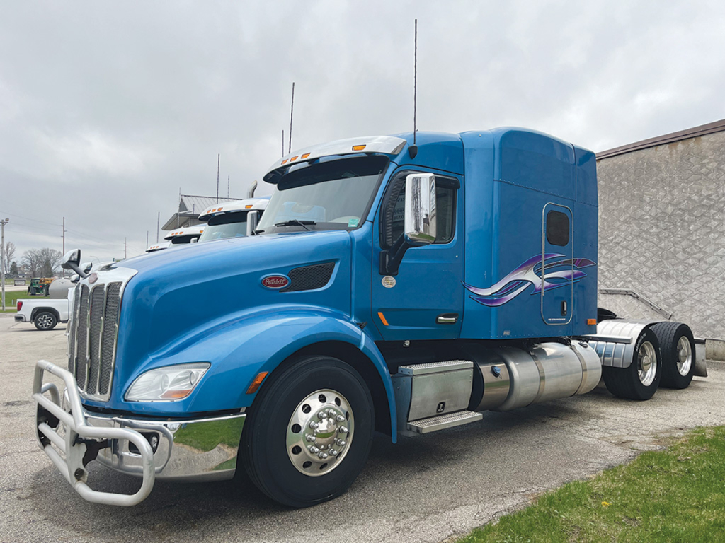 Peterbilt 579 UltraCab tractor in blue with aerodynamic fairing and chrome bumper guard. Side fuel tanks, sleeper cab, and rear wheels visible.