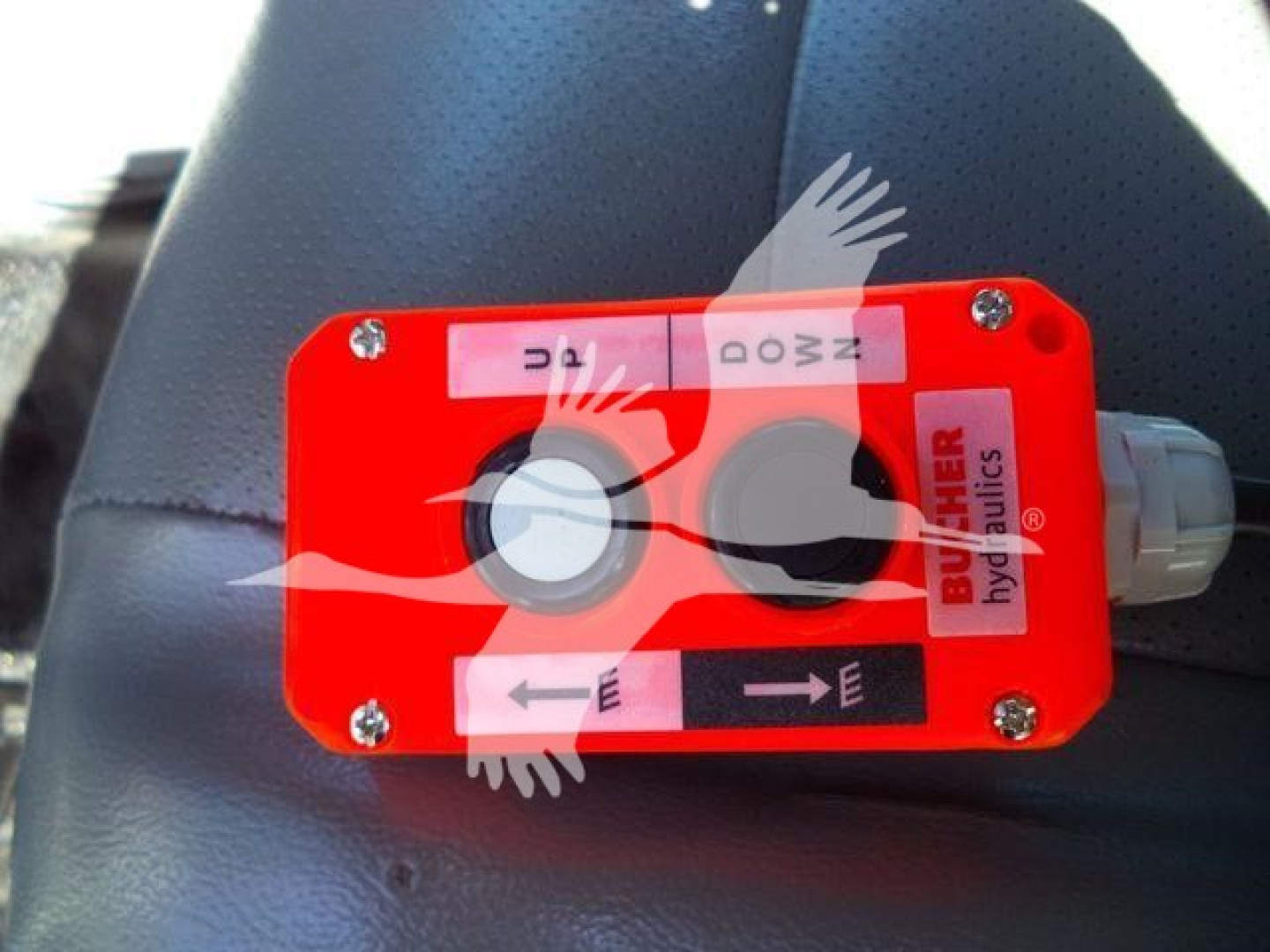 Red hydraulic valve block with white labels and arrows on a blue vehicle seat.