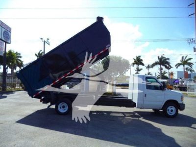 Truck bed tilted dump body on a white Ford E-450 chassis cab dump truck, mid-angled view showing rear wheels and hydraulic elevation.