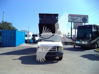 Ford E450 dump truck front view with raised dump bed, blue portable shed left, bus and sign in background.