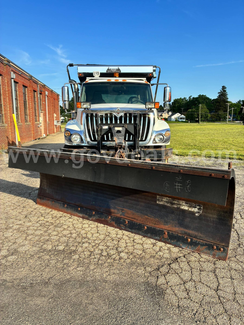 Truck front view with snow plow attachment, International 7400 dump truck, chrome grille, large headlights, side mirrors, hood ornament, and plow blade in foreground.