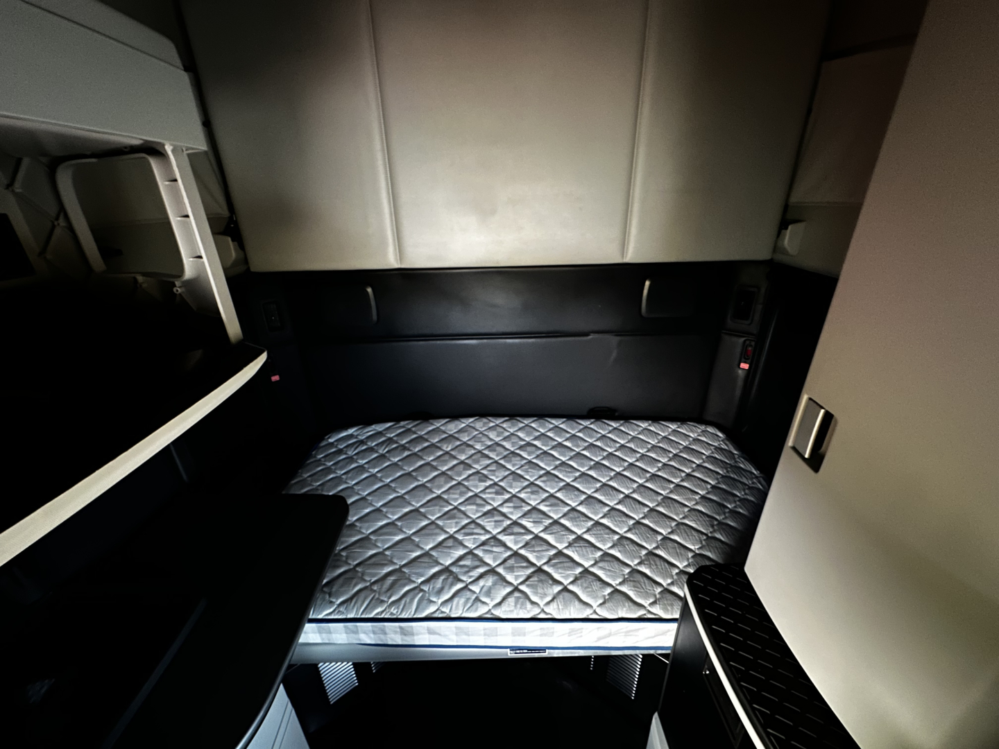 Truck cab sleeper interior showing a quilted mattress in the bunk area of a Kenworth T680 sleeper with surrounding panels and storage compartments.