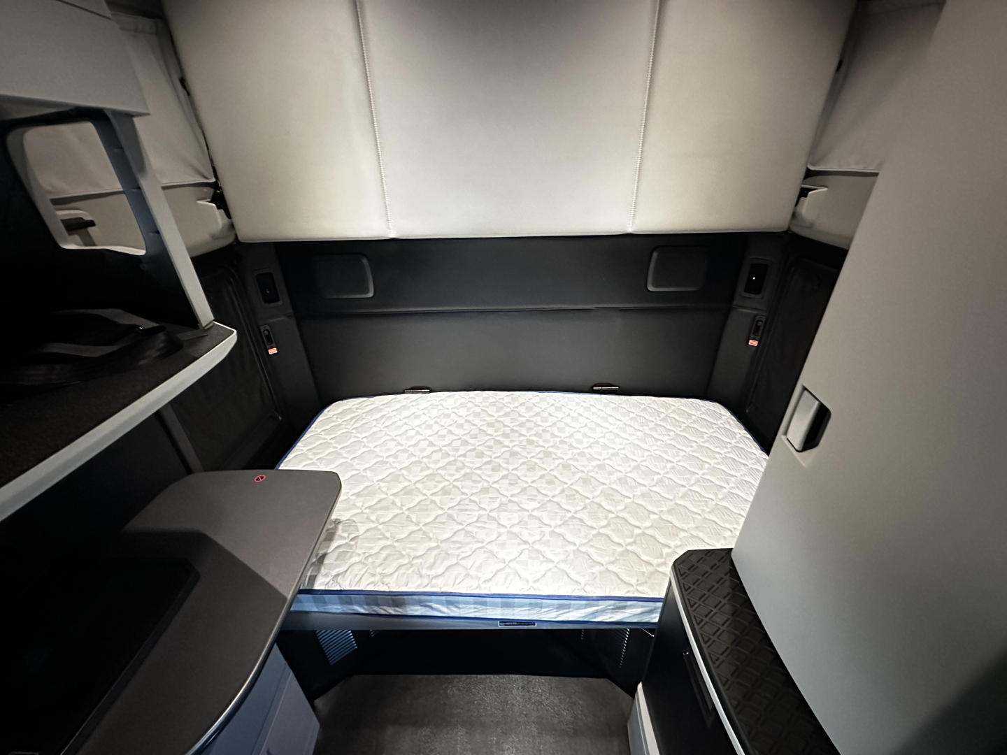 Kenworth T680 sleeper cab interior showing rear bed mattress in living area with cabinets and side panels.