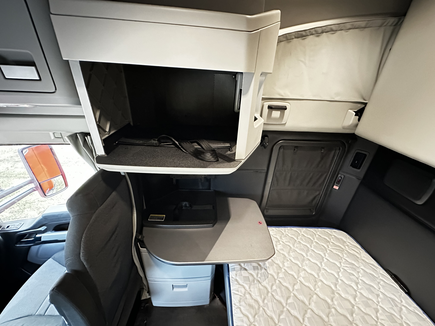 Kenworth T680 sleeper cab interior showing open overhead storage with a folded table and small shelf, and a compact desk area by the bed.