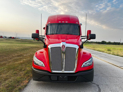 Front view of a red 2023 Kenworth T680 truck with chrome grille, black bumper, and side mirrors, in a parking area.