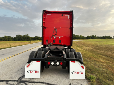 Kenworth T680 rear view showing red sleeper cab, dual rear axles, mud flaps with MHC logo, on a rural road.