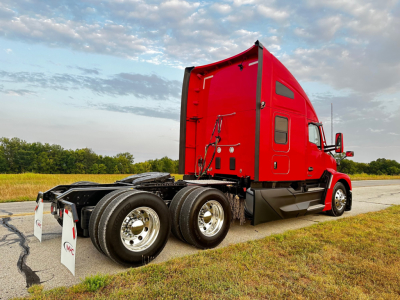 Kenworth T680 tractor with red sleeper cab, dual rear axles, and chrome wheels visible in a roadside setting.