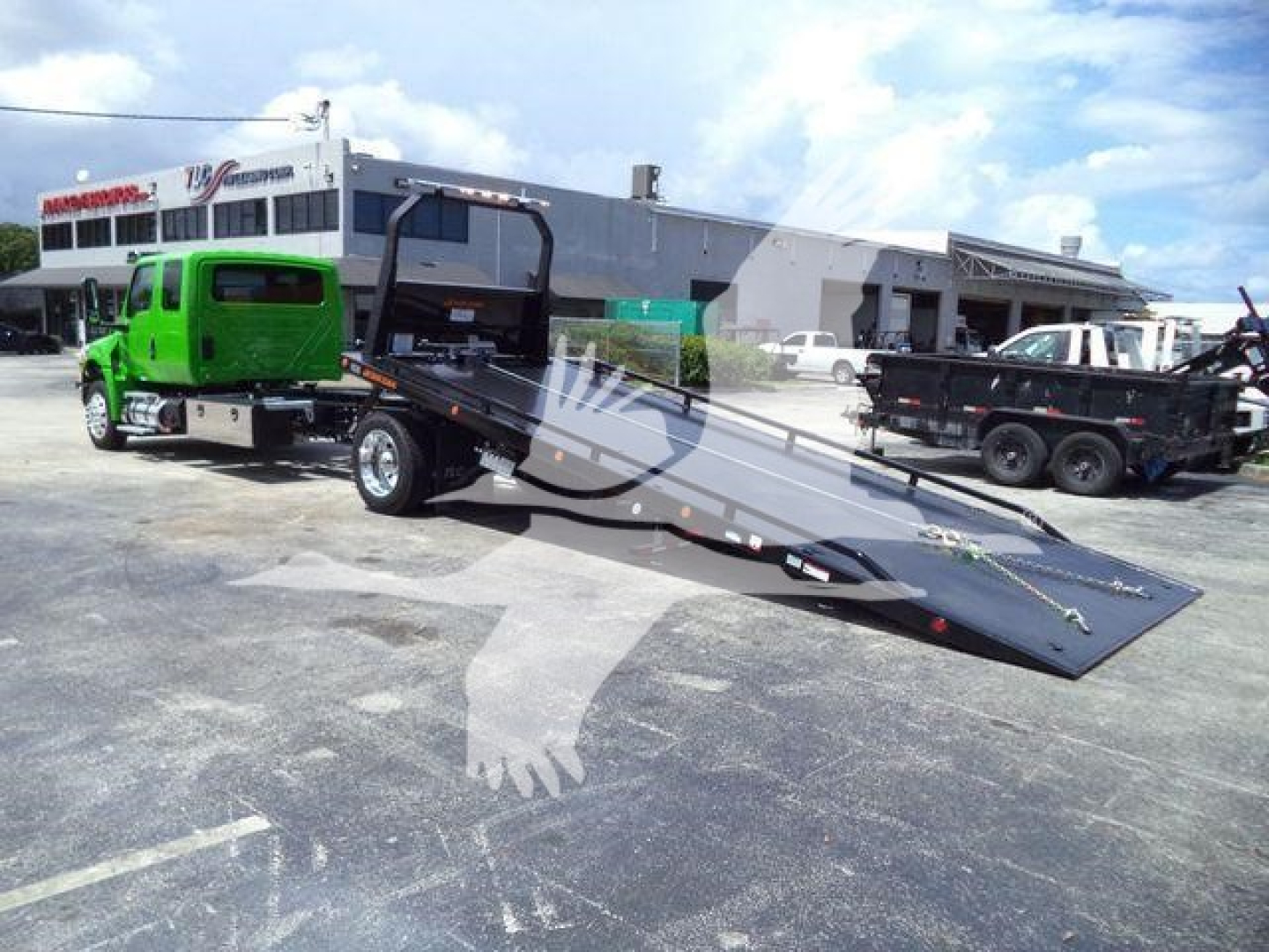 Tow truck with a green cab and a flatbed rollback trailer in a dealership lot, 2027 International MV, visible text on building and vehicles.