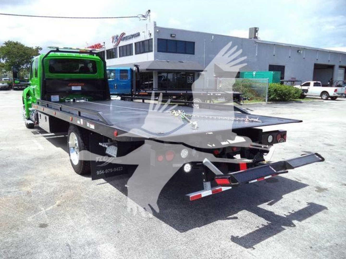 Flatbed tow truck in a dealership lot showing green cab and black steel flatbed; rear ramp and hitch, cargo area with side lights, building in background.
