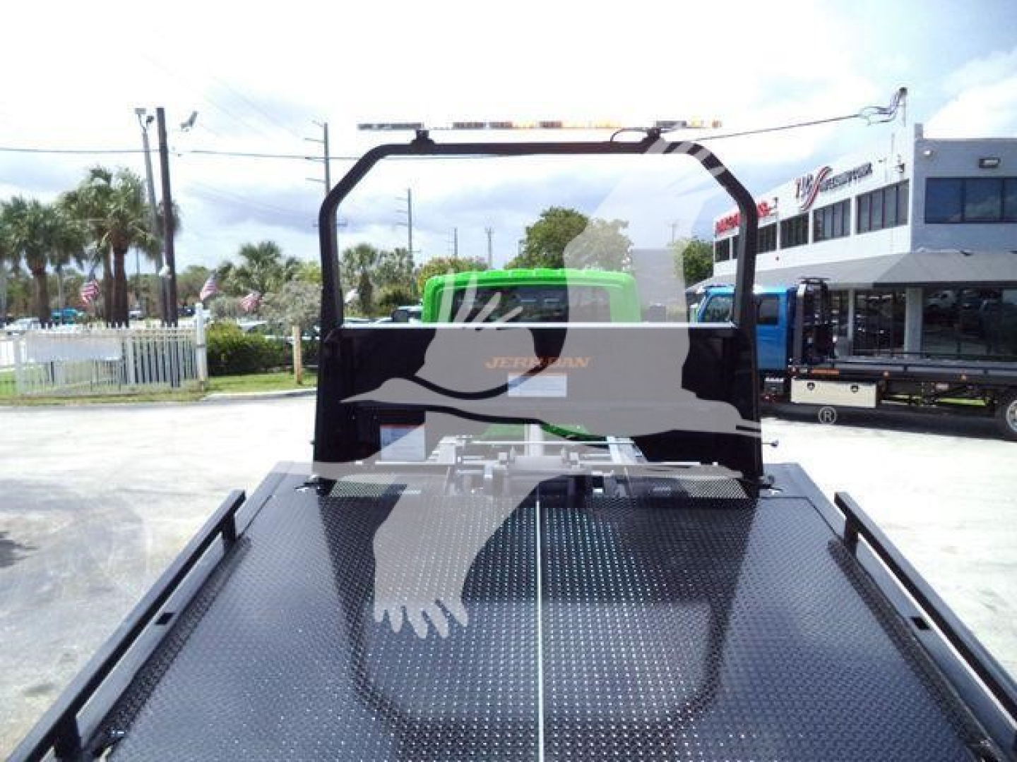 Truck bed with a black flatbed and a green cab visible in background, roll-back equipment, chain link fence and dealership in distance.