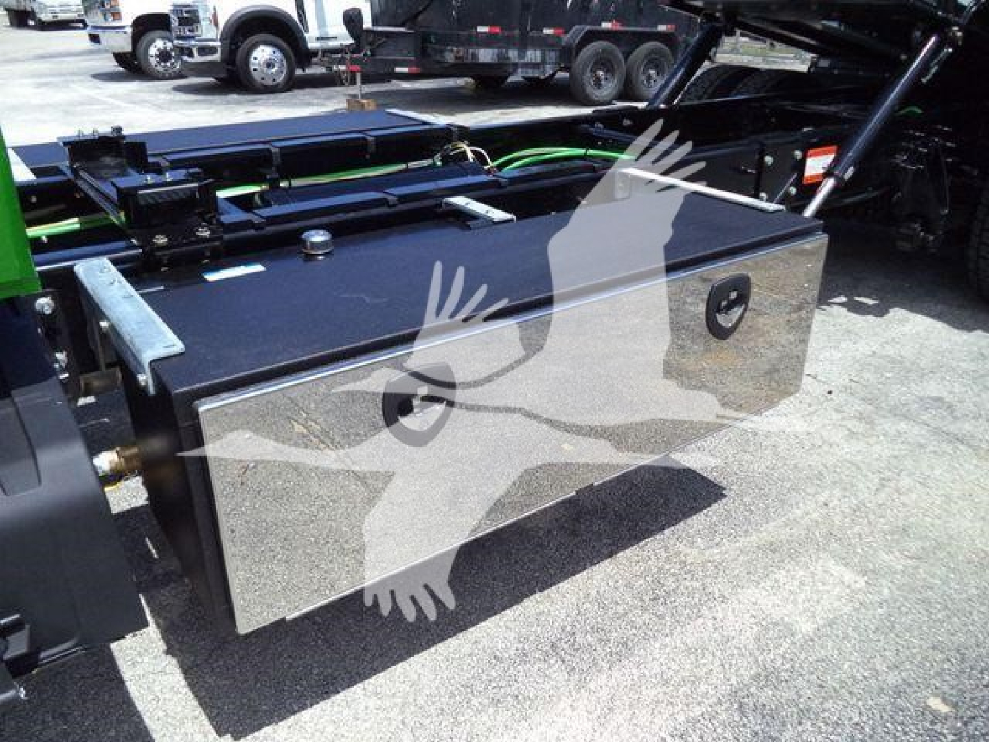 Truck toolbox mounted on side of truck frame with metal silver finish, hinged lid, and recessed handles; surrounding chassis and fuel lines visible.