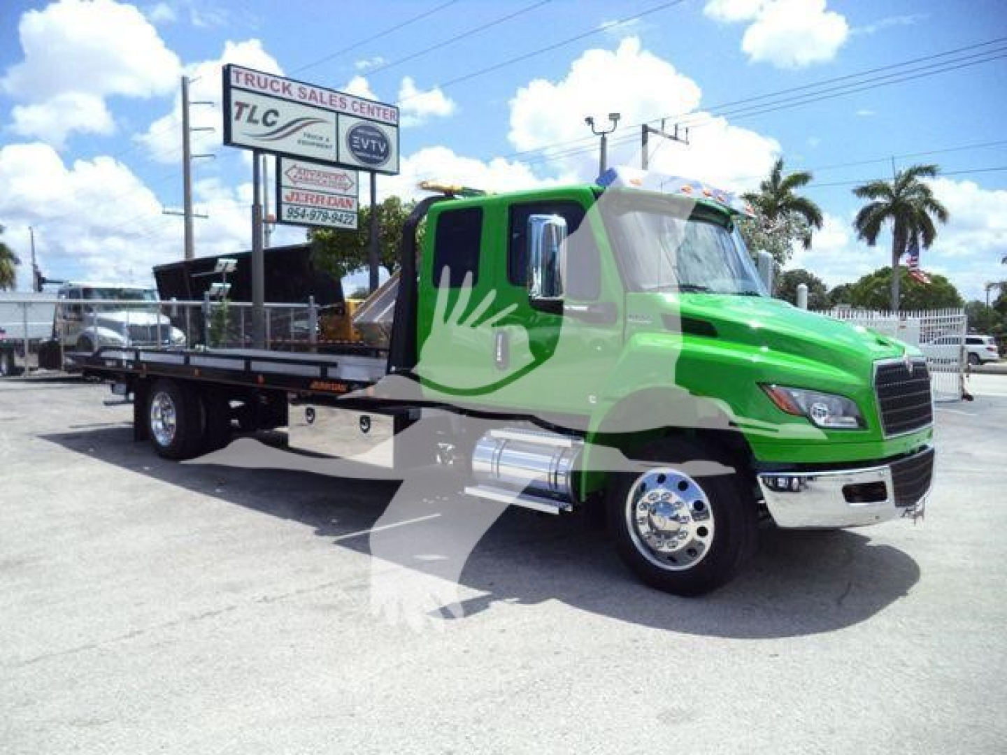 Green International MV rollback truck viewed from side-front angle, with extended flatbed, chrome exhaust, and black chassis; TLC sign in background.