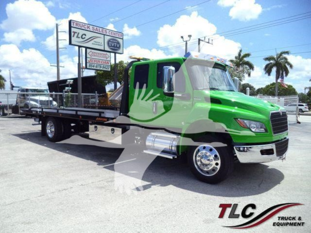 Green International MV rollback truck with flatbed in a dealership lot, stock number J24111, TLC sign in background.