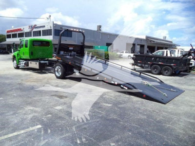 Tow truck with a green cab and a flatbed rollback trailer in a dealership lot, 2027 International MV, visible text on building and vehicles.