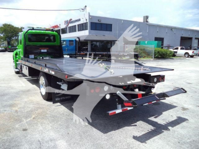 Flatbed tow truck in a dealership lot showing green cab and black steel flatbed; rear ramp and hitch, cargo area with side lights, building in background.