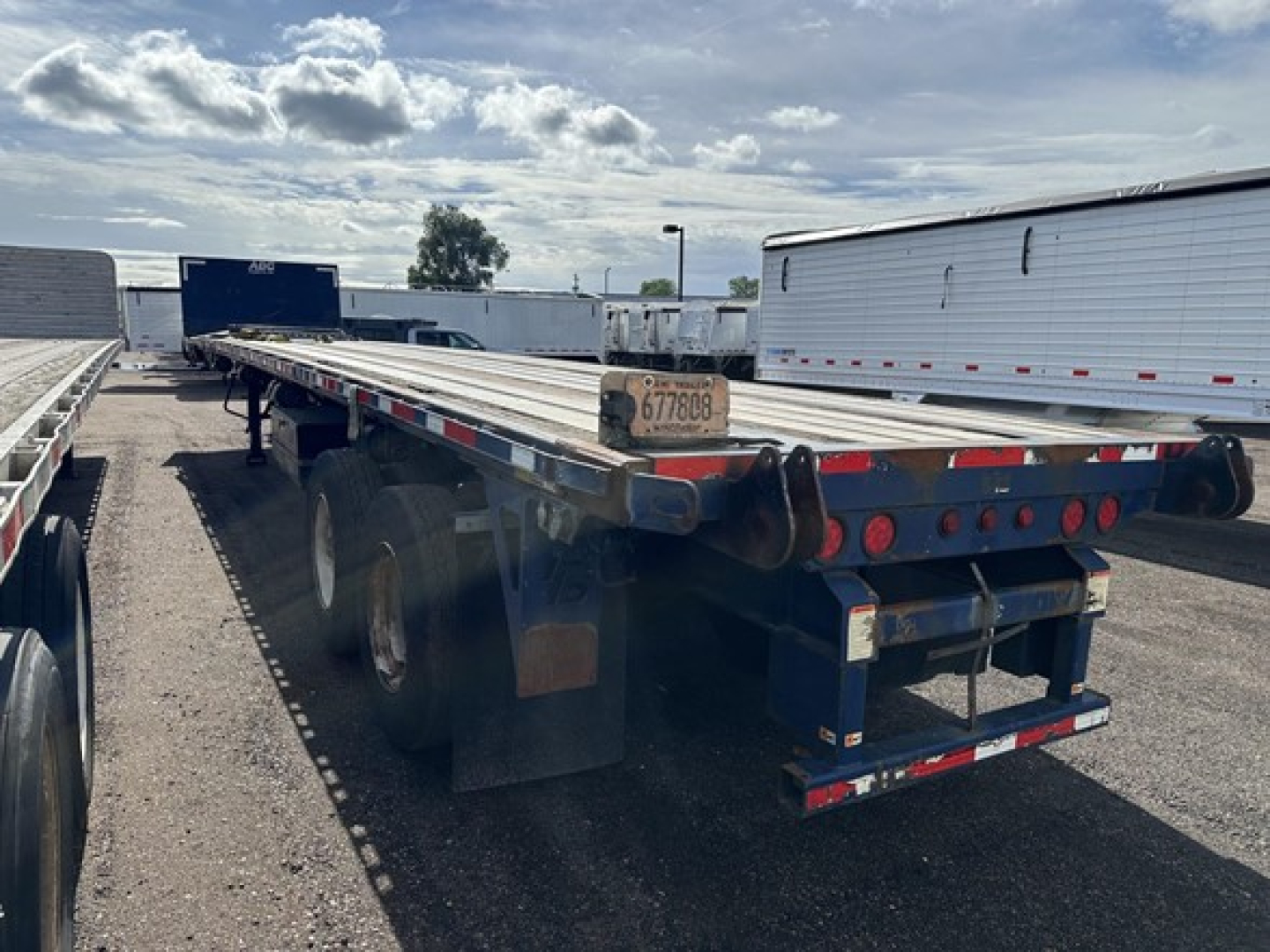 Flatbed trailer rear and side view showing tandem axles and rear lights with a license plate visible.