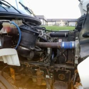 Engine bay of a Freightliner M2 106 showing various hoses, wiring, and components; blue hose with clamp, coolant reservoir, exposed engine and frame hardware.