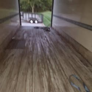 Interior view of a trailer with wooden floor and ramp at back, showing a trailer interior and a small trailer outside, with some straps on the floor.