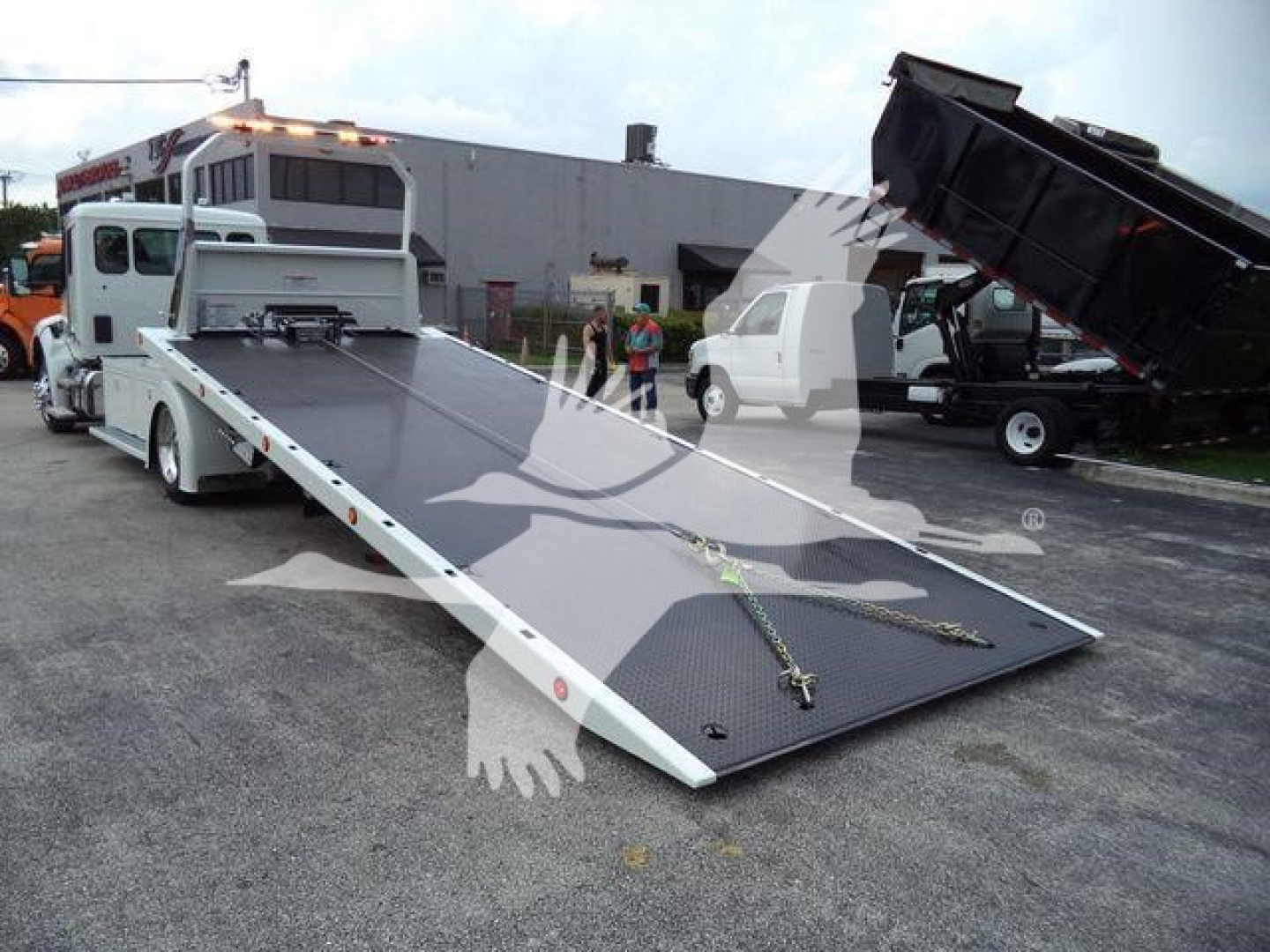 Kenworth flatbed rollback truck with a lowered ramp at a dealership lot showing a white truck in background.