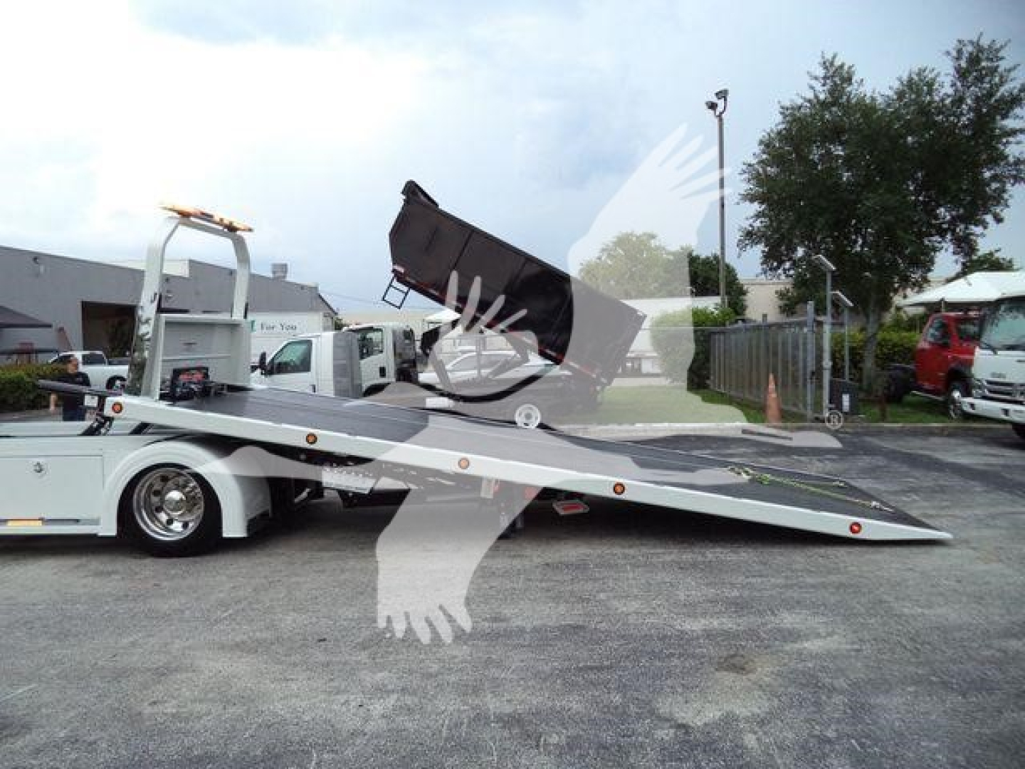 Kenworth truck in a rollback tow setup with a lowered tilt bed and a black toolbox on the bed; background shows a parking lot and other vehicles.