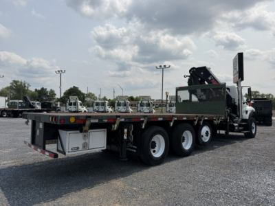 Flatbed crane truck with triple rear axles and crane arm in folded position. Side panels and rear reflectors visible; no legible text on the truck from this view.