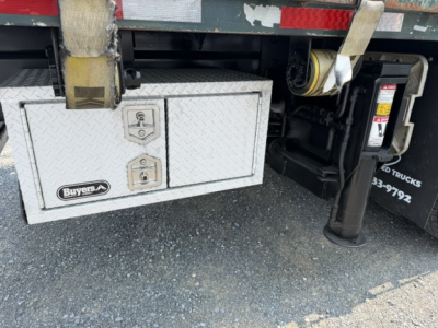 Truck undercarriage showing a diamond-plated tool/storage box with two latches and a Babys-A badge, hydraulic stabilizer leg visible, gravel surface.