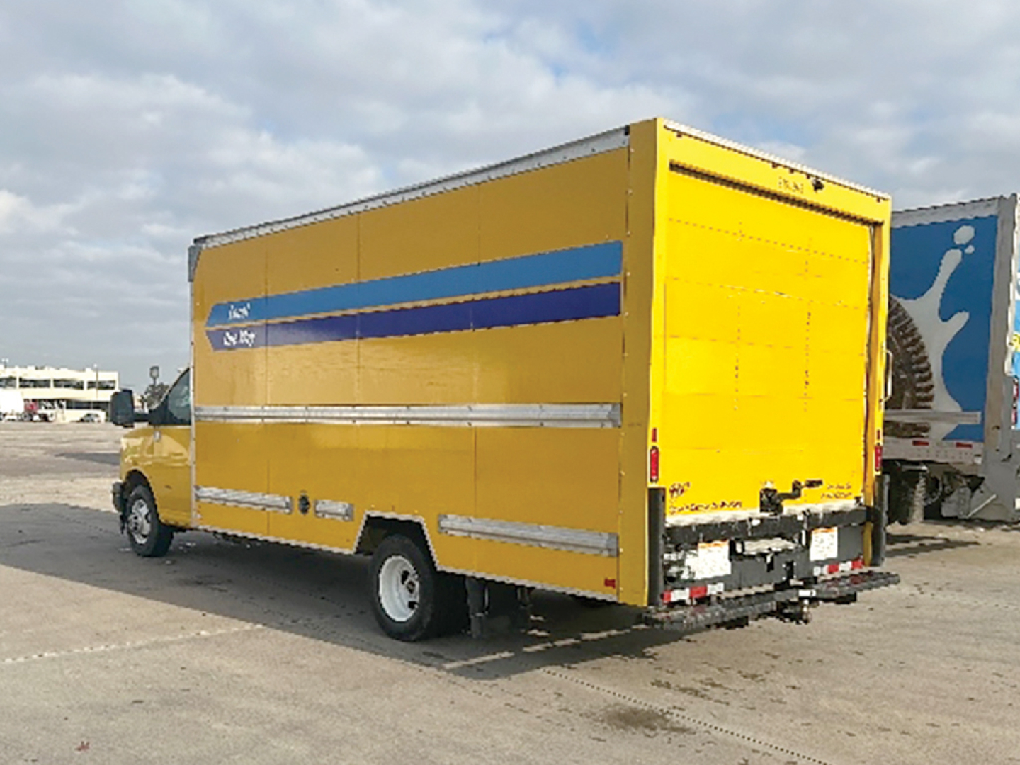 GMC Savana cargo truck with yellow box and blue stripe viewed from rear-left side.