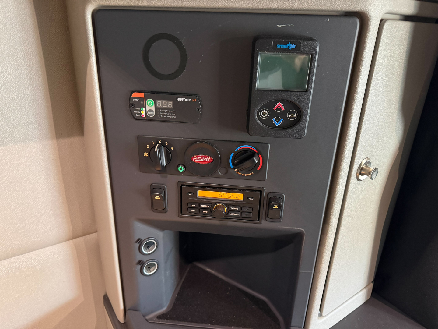 Truck cab interior panel showing controls: circular gauge, digital display, climate knobs, radio, and storage pocket.