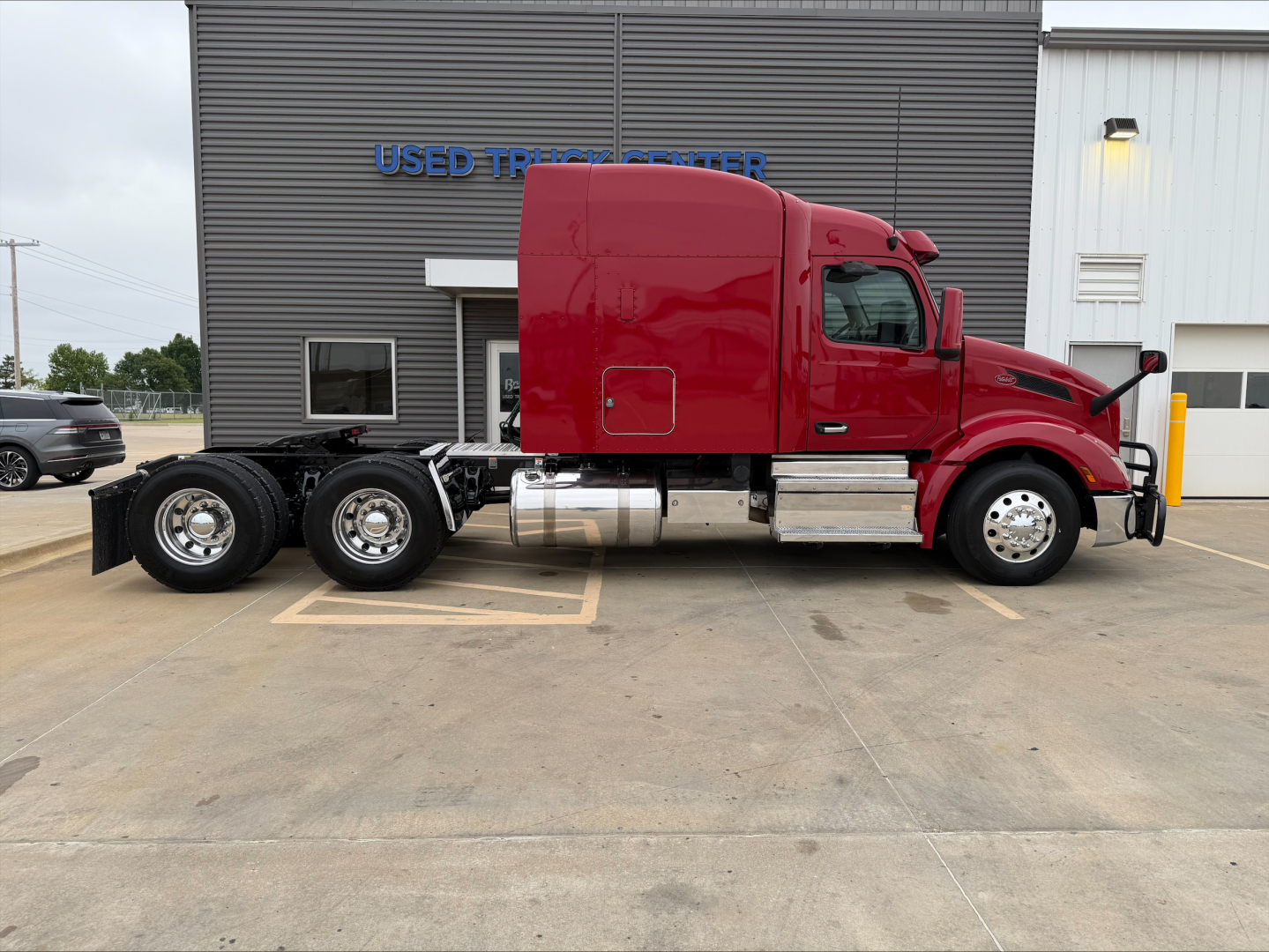 Peterbilt 579 tractor viewed from side, red cab, black chassis, dual rear axles, chrome fuel tanks, parked in front of a building with 'USED TRUCK CENTER' sign.