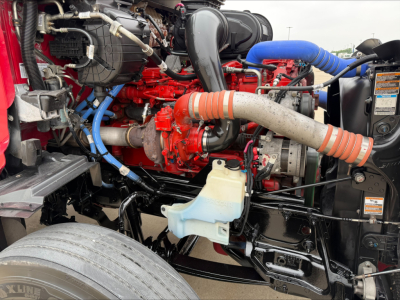 Peterbilt 579 engine bay showing red engine block, blue coolant lines, orange intake hose, black intercooler piping, windshield washer reservoir, and various hoses.
