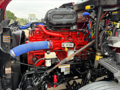 Peterbilt 579 engine bay showing Cummins X15 engine with red intake manifold and blue intercooler hose, visible fluids reservoirs and wiring harnesses.
