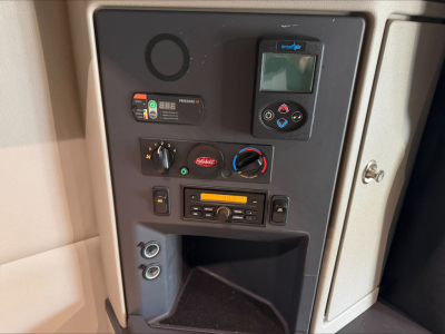 Truck cab interior panel showing controls: circular gauge, digital display, climate knobs, radio, and storage pocket.