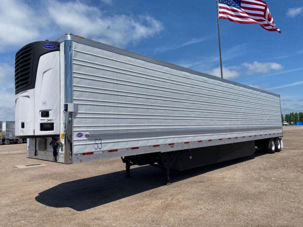 Utility reefer trailer with a mounted refrigeration unit at the front, silver corrugated sides, undercarriage visible, American flag in background.